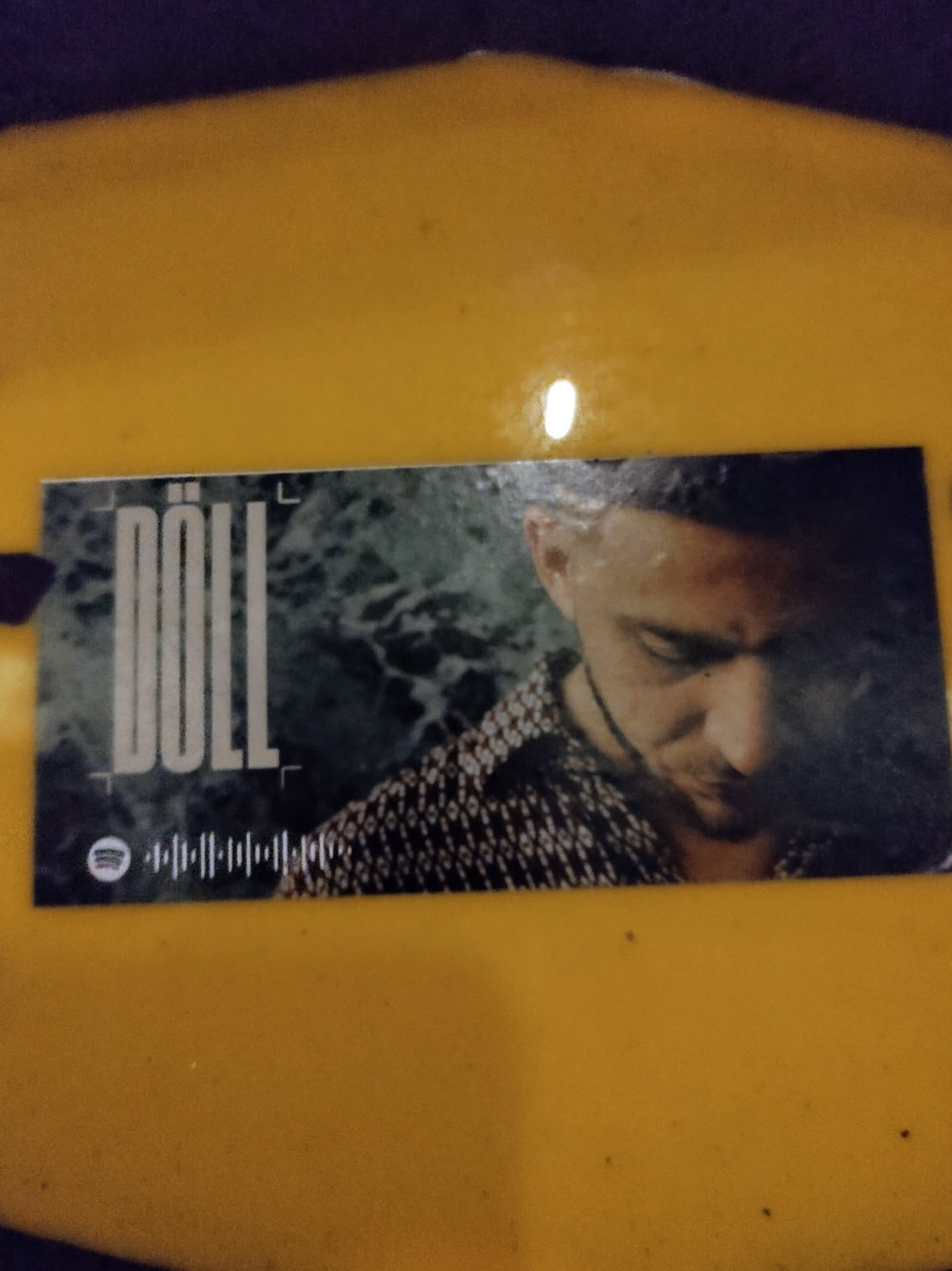 This sticker features a grayscale image of a man with short hair, wearing a patterned shirt.  The word "DÖLL" is prominently displayed in a stylized font.  There is also a Spotify code visible at the bottom.