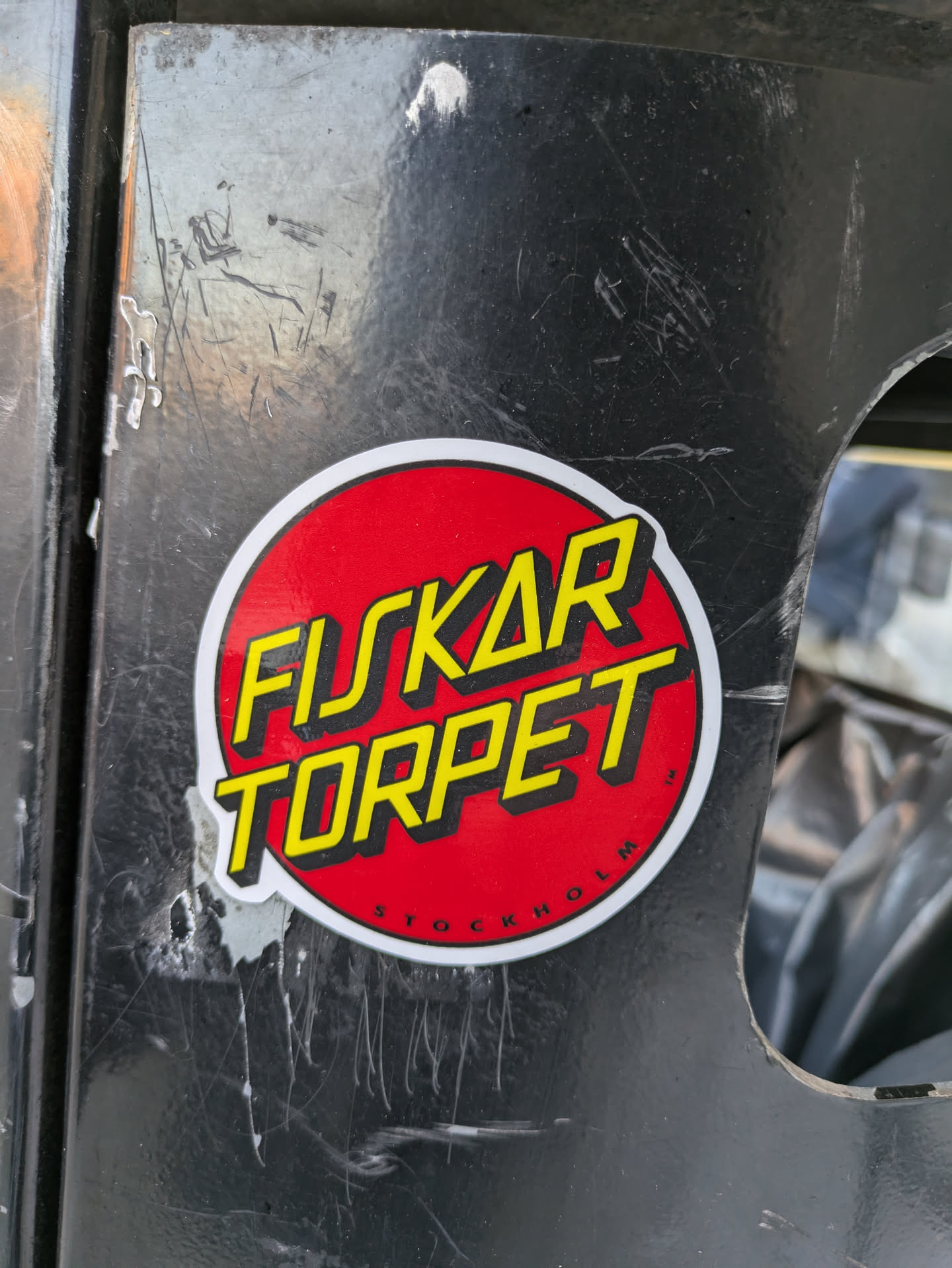 A red and yellow round sticker with the words "FUSKAR TORPET" written in a stylized font.  "STOCKHOLM" is written in smaller text at the bottom. The sticker is stuck on a dark grey surface. 
