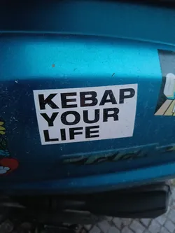 A sticker with the text "KEBAP YOUR LIFE" in black on a white background, affixed to a teal-colored vehicle. The sticker has a simple, bold design.