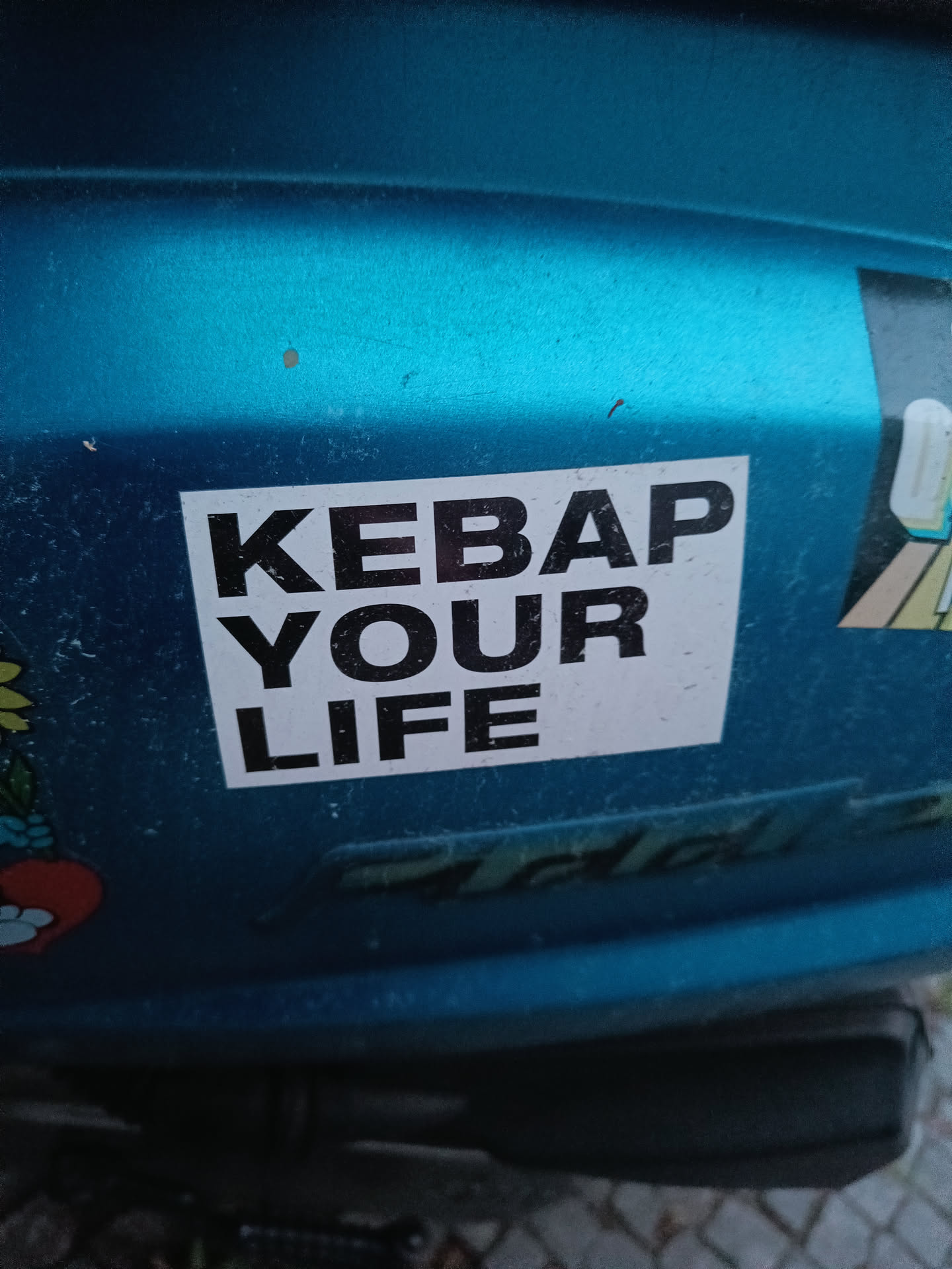 A sticker with the text "KEBAP YOUR LIFE" in black on a white background, affixed to a teal-colored vehicle. The sticker has a simple, bold design.
