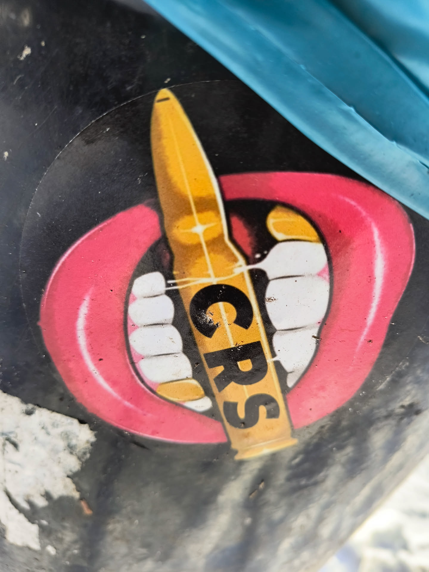 The image shows a round sticker with a black background. The sticker features a red-lipped mouth with white teeth, and a bullet is used as one of the teeth. The letters CRS are printed on the bullet.
