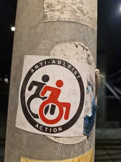 Street sticker A square paper sticker stuck onto a metal pole. It features a circular design parodying the Antifascist Action logo, enclosed in a thick black ring containing the text 'ANTI-ABLEIST' along the top and 'ACTION' along the bottom. Inside the circle are two active wheelchair pictogram figures moving forward, one in black on the left and overlapping with another in red in the foreground.