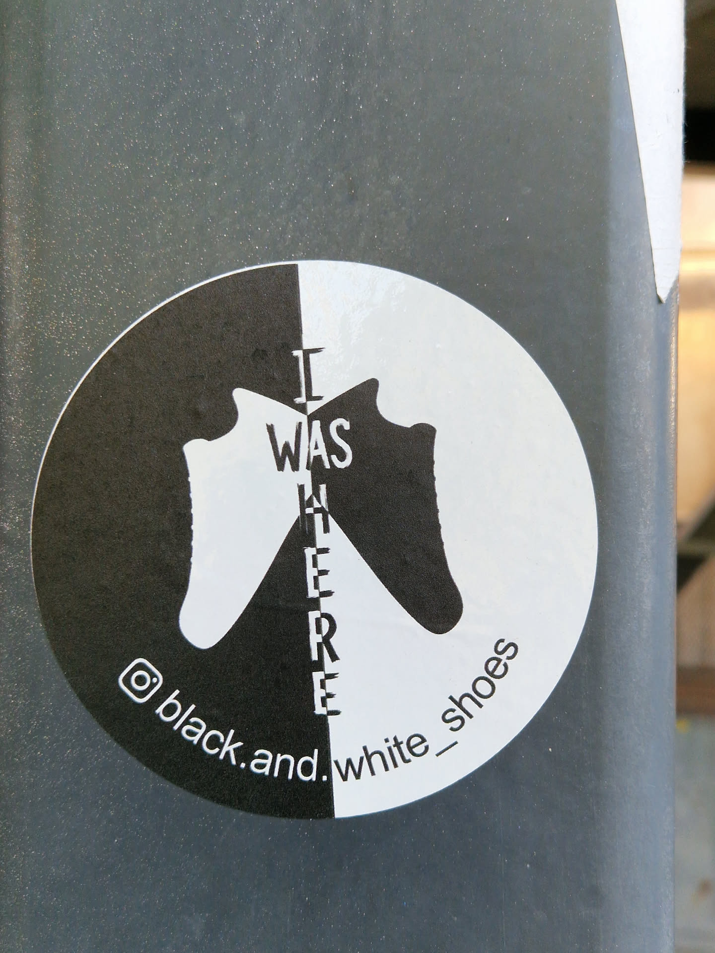 A circular sticker divided vertically into black and white halves. The left black half features a white silhouette of a shoe facing inward, while the right white half displays a black silhouette of a shoe also facing inward. The shoes are symmetrical, meeting in the middle. Vertically centered between the shoes, in a contrasting color (white on black, black on white), is the text 'I WAS HERE'. Along the bottom curve of the sticker, an Instagram logo precedes the handle 'black.and.white_shoes', written in white on the black side and black on the white side.