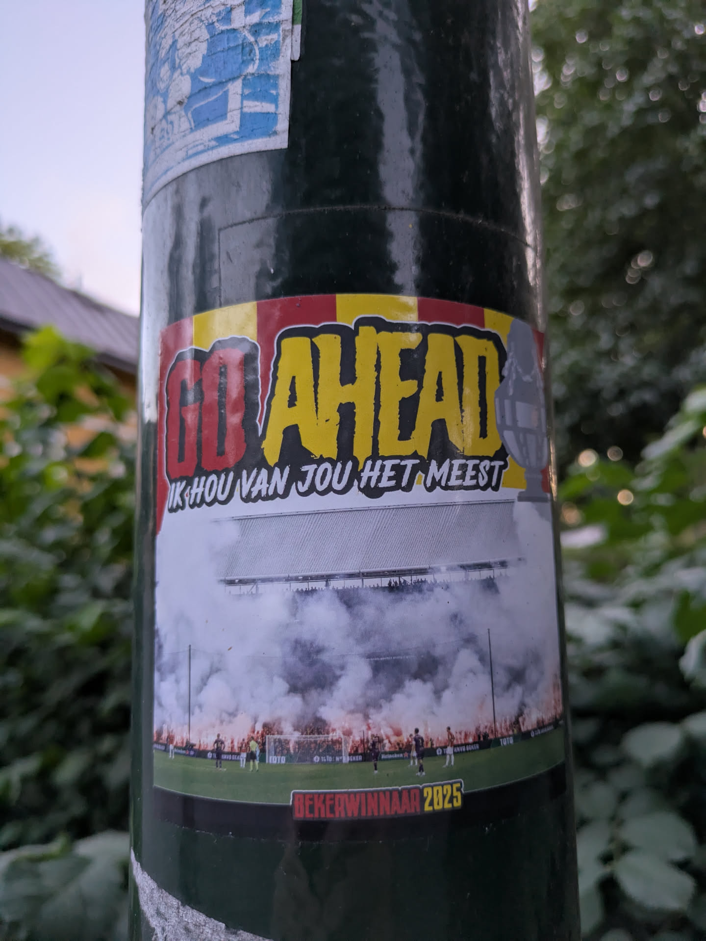 The sticker is rectangular with slightly rounded corners, adhered to a dark green pole. The top portion features a banner with red and yellow stripes, reminiscent of a flag. Prominently displayed are the words "GO AHEAD" in a bold, stylized font; "GO" is red, and "AHEAD" is yellow, both outlined in black and white. To the right of "AHEAD" is a grey, stylized trophy cup. Below "GO AHEAD" in smaller white text is the Dutch phrase "IK HOU VAN JOU HET MEEST" (I love you the most). The central and largest part of the sticker depicts a vibrant football stadium scene, likely during a night match. The stands are visible with spectators, and the field below is engulfed in thick white smoke from numerous red flares or pyrotechnics lit along the lower stands. Football players are visible on the pitch. Small, partially obscured logos like "TOTO", "Heineken", and "KNVB BEKER" can be seen within the stadium image. The bottom of the sticker has a black band with red text that reads "BEKERWINNAAR 2025" (Cup Winner 2025), indicating a future aspiration.