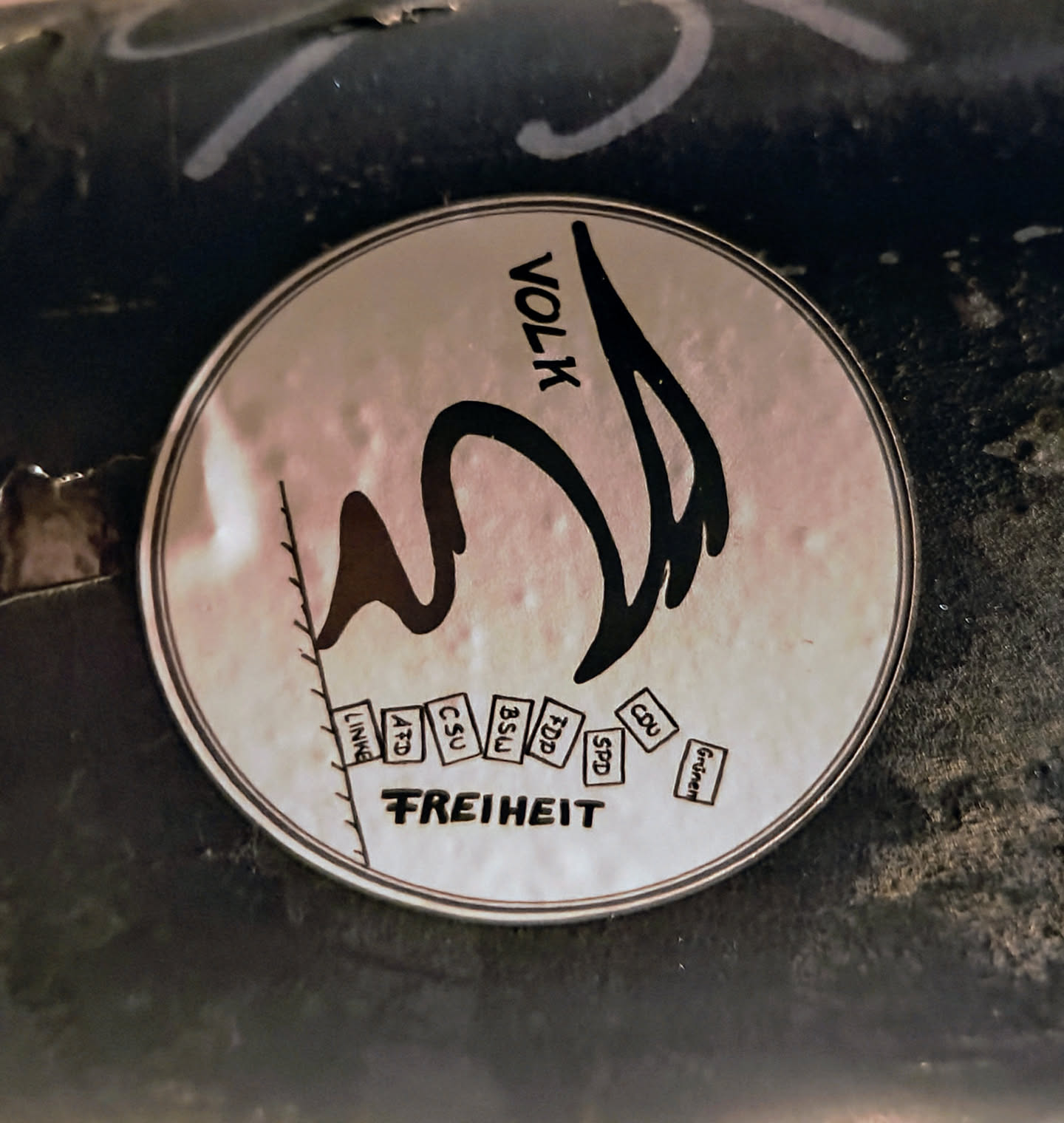 Round sticker with a drawing of a bird and the word VOLK above it. Below the bird are small boxes with German political party names: LINKE, AFD, CSU, BSU, FDP, SPD, CDU, and GRÜNE. At the bottom, the word FREIHEIT (freedom) is written. The sticker seems to be a political statement.