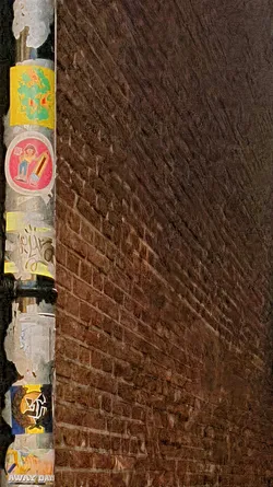 The image features a pole covered with various stickers set against a backdrop of a brick wall. The stickers are colorful and feature different designs. One sticker depicts a figure with a pencil and appears to warn against something, due to the red circle and bar over the image.