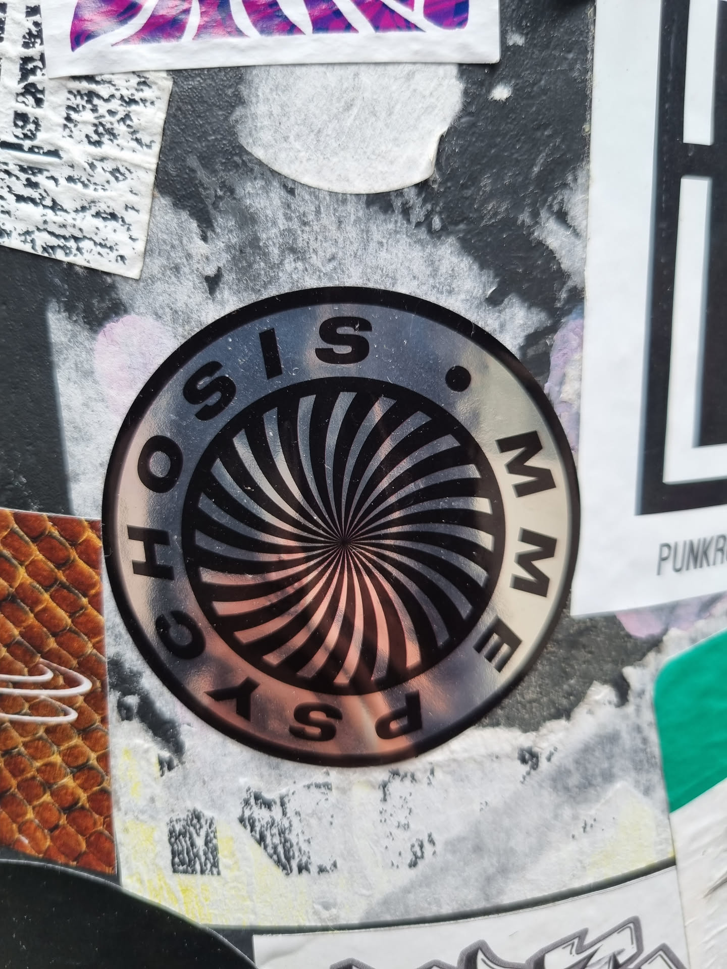 A round, silver sticker with a black and white hypnotic spiral design. The words \"PSYCHOSIS MEME\" are written around the spiral in a circular pattern. The sticker appears to be affixed to a surface with other stickers and graffiti.