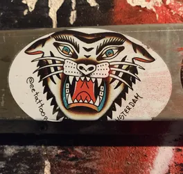 Oval sticker featuring a traditional tattoo style tiger head with its mouth open, showing sharp teeth.  The tiger's eyes are a striking blue. The background is white, and the text '@eetattoos' and 'AMSTERDAM' are visible.