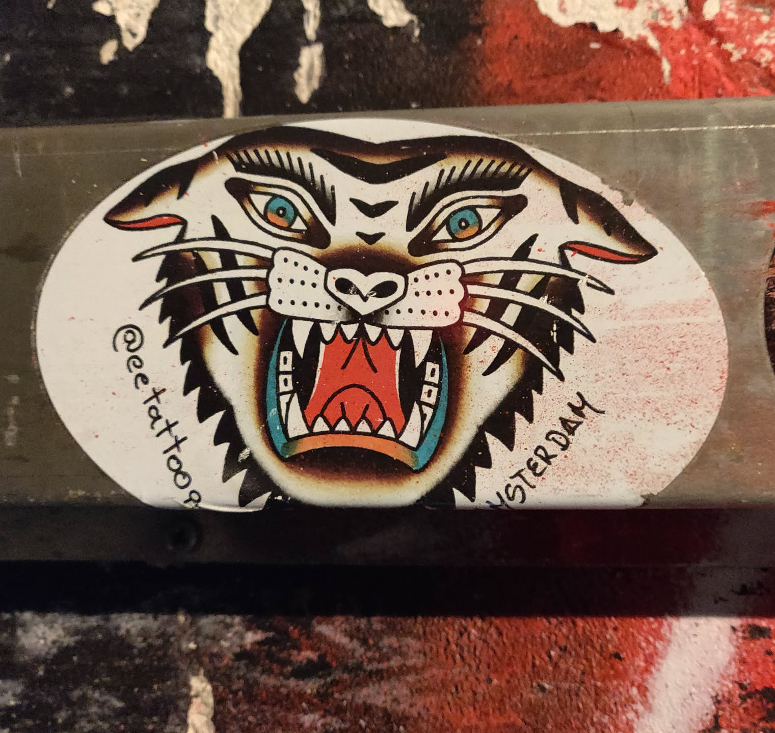 Oval sticker featuring a traditional tattoo style tiger head with its mouth open, showing sharp teeth.  The tiger's eyes are a striking blue. The background is white, and the text '@eetattoos' and 'AMSTERDAM' are visible.