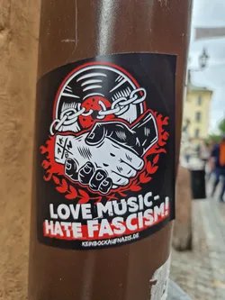 A sticker with a design showing two hands shaking, bound by a chain to a vinyl record. Flames are depicted behind the hands. The text on the sticker reads: "LOVE MUSIC- HATE FASCISM!" and includes a website address: keinbockaufnazis.de