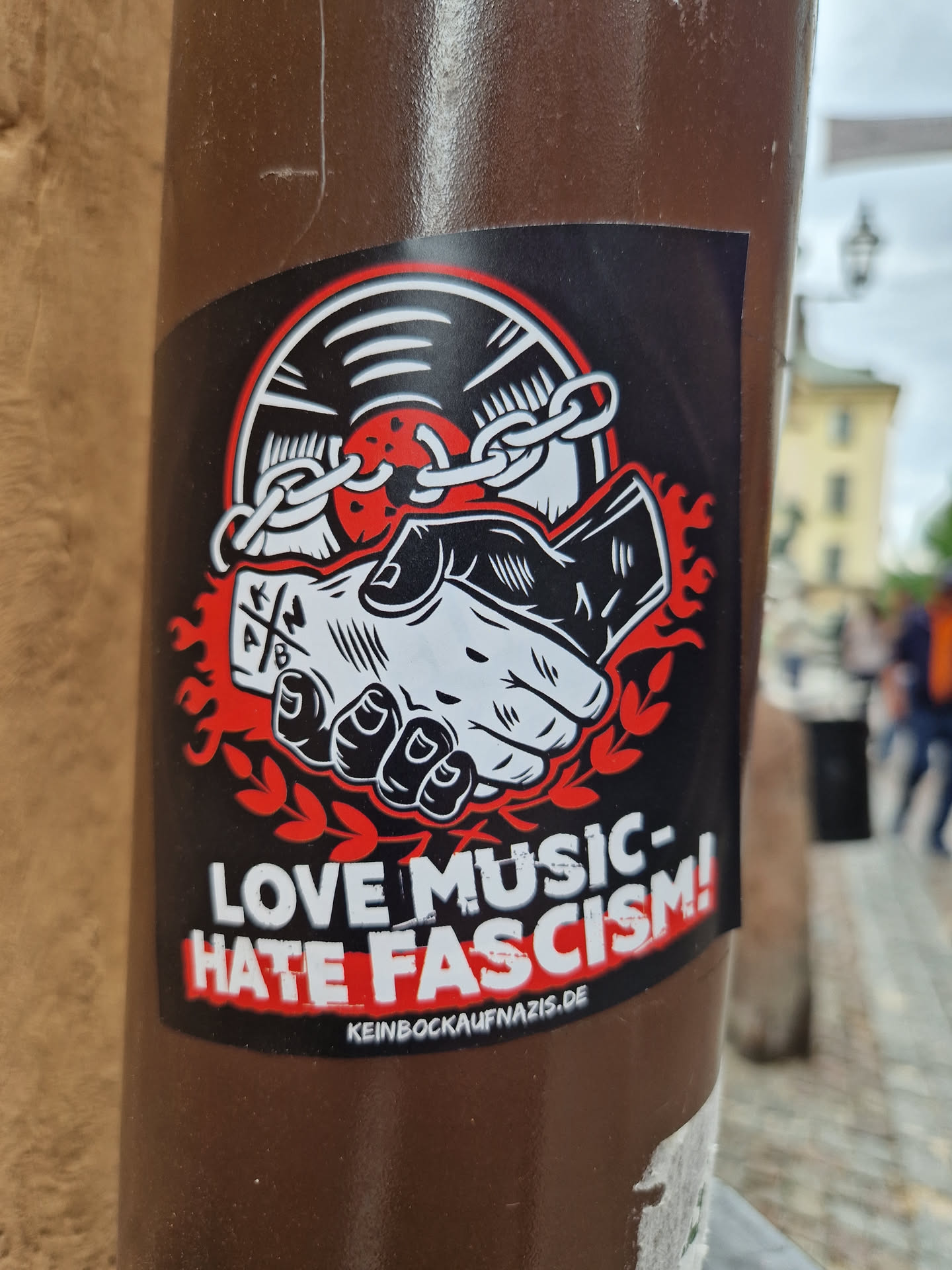 A sticker with a design showing two hands shaking, bound by a chain to a vinyl record. Flames are depicted behind the hands. The text on the sticker reads: "LOVE MUSIC- HATE FASCISM!" and includes a website address: keinbockaufnazis.de