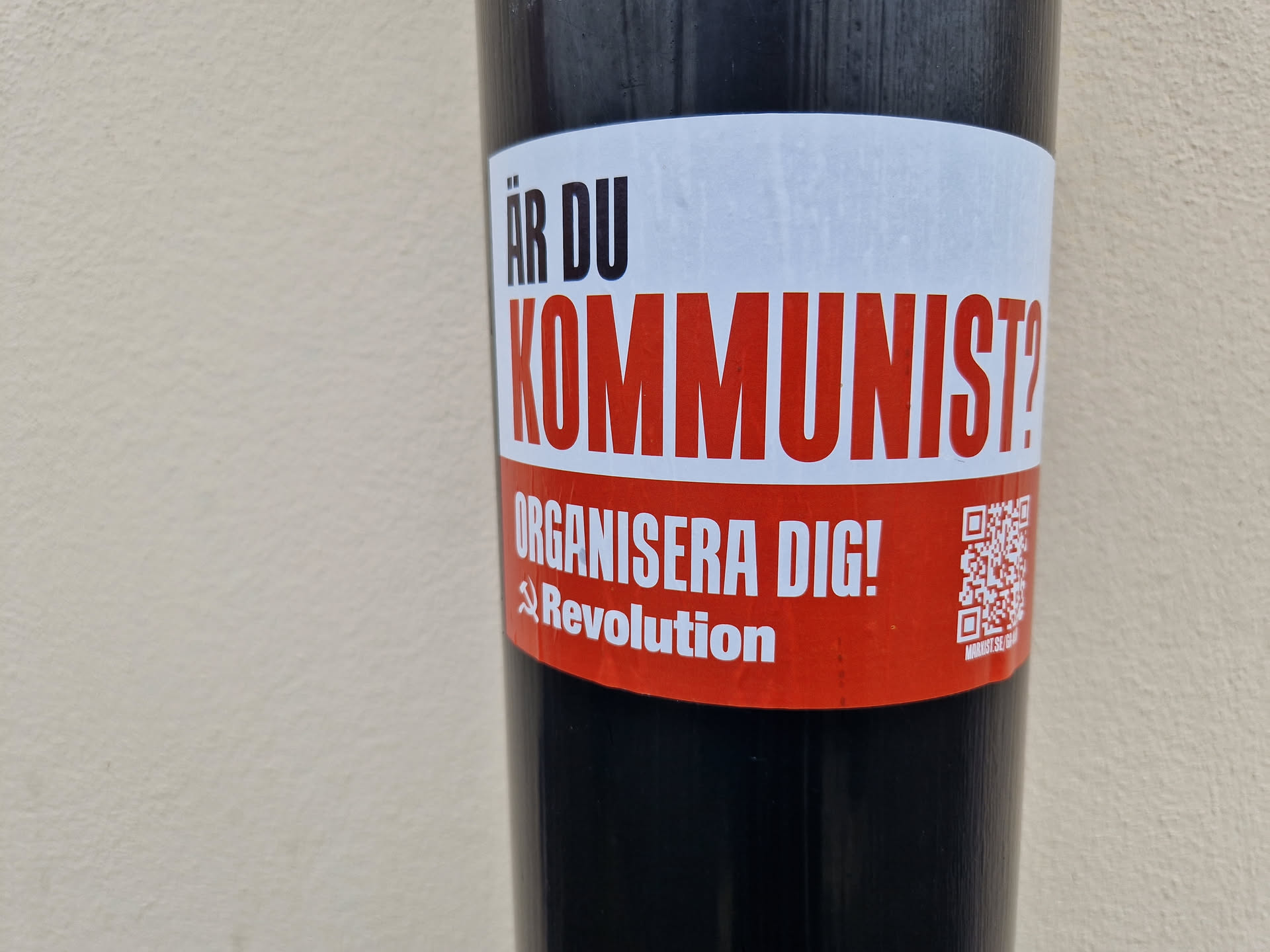 A red and white sticker with the text "Är du kommunist?" (Are you a communist?) and "Organisera dig!" (Organize yourself!). It also features a QR code and the word "Revolution" with a hammer and sickle symbol.