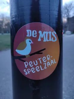 A round sticker with an illustration of a bird on a branch.  The text on the sticker says "De Mus Peuter-Speelzaal".