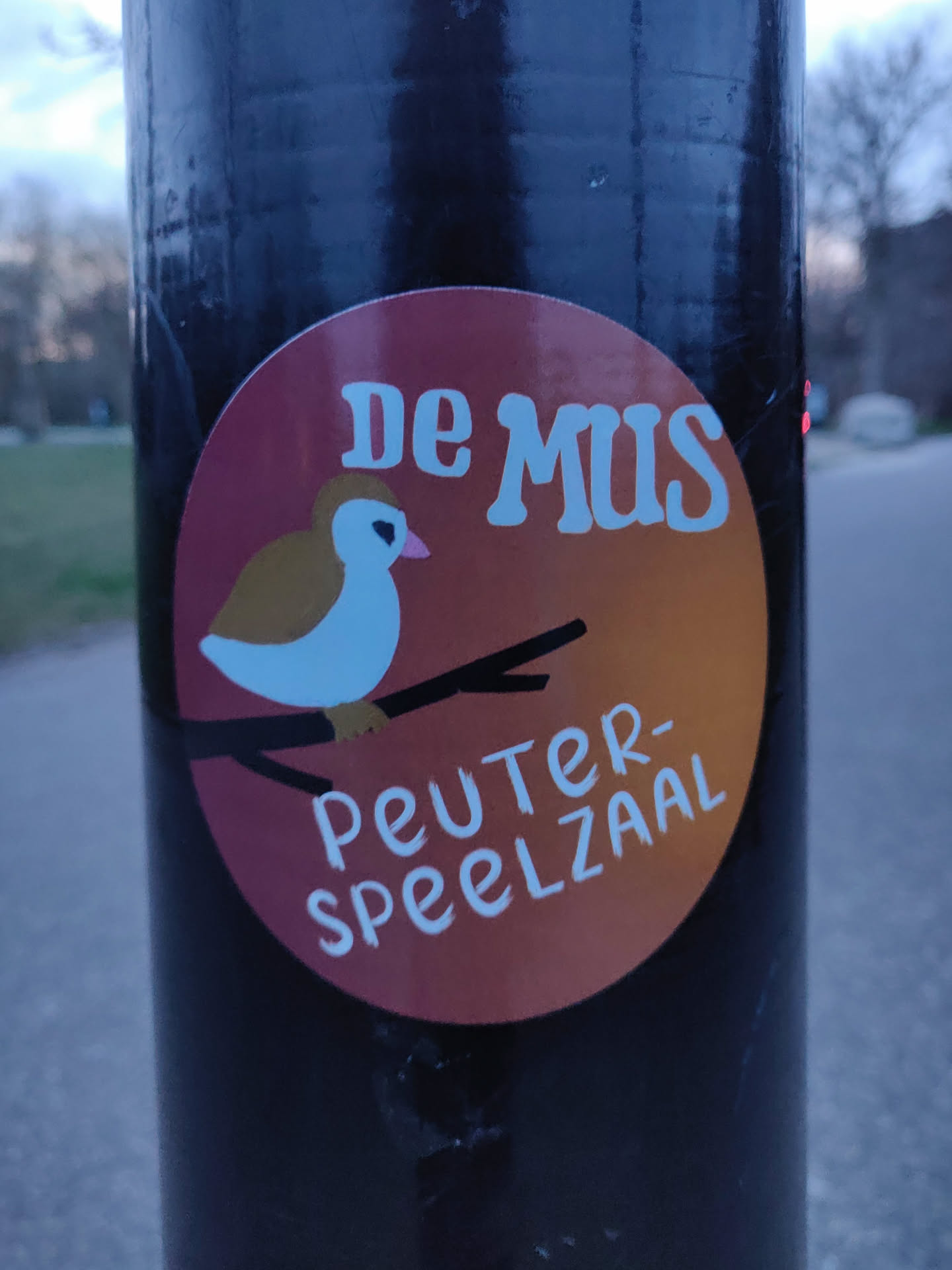 A round sticker with an illustration of a bird on a branch.  The text on the sticker says "De Mus Peuter-Speelzaal".