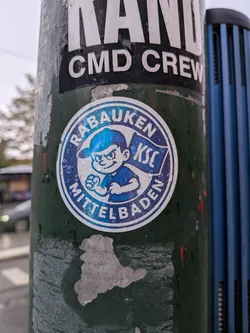 Street sticker A circular sticker on a green pole covered with light water droplets. The sticker has a white outer border followed by a blue circular band containing the bold white text 'RABAUKEN' along the top arc and 'MITTELBADEN' along the bottom arc. Inside the circle is a cartoon boy wearing a blue cap turned backward, posing determinedly with clenched fists. Behind him waves a blue flag bearing the letters 'KSC' in white, referencing Karlsruher SC.