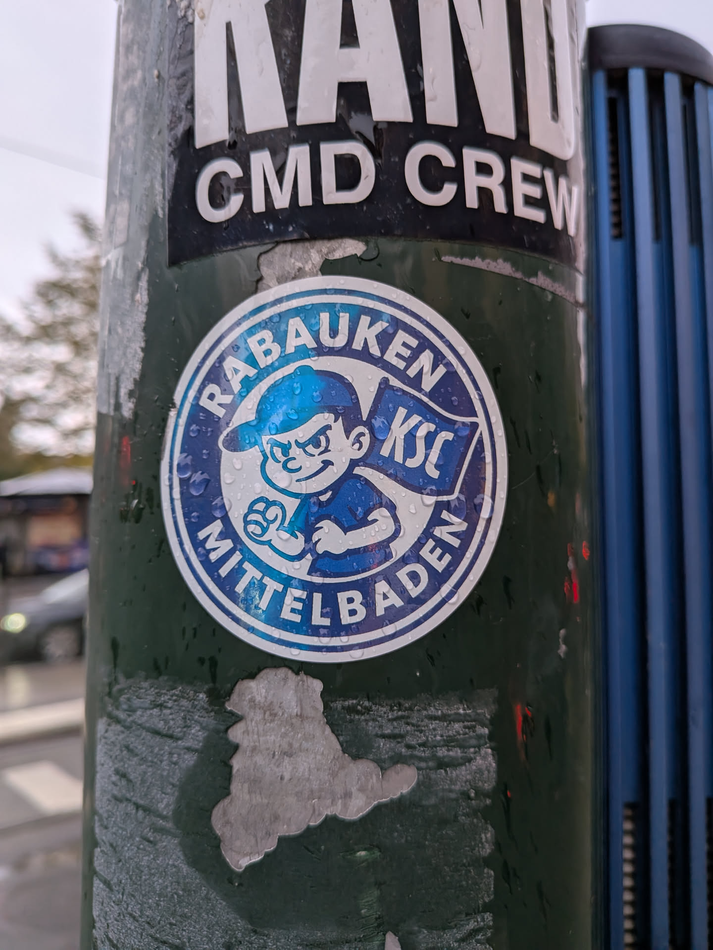 A circular sticker on a green pole covered with light water droplets. The sticker has a white outer border followed by a blue circular band containing the bold white text 'RABAUKEN' along the top arc and 'MITTELBADEN' along the bottom arc. Inside the circle is a cartoon boy wearing a blue cap turned backward, posing determinedly with clenched fists. Behind him waves a blue flag bearing the letters 'KSC' in white, referencing Karlsruher SC.