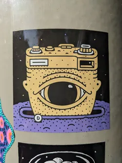 Sticker of a camera with one eye, set in space. The camera is yellow and the background is black with purple accents. There is a small letter D in a circle on the purple part.