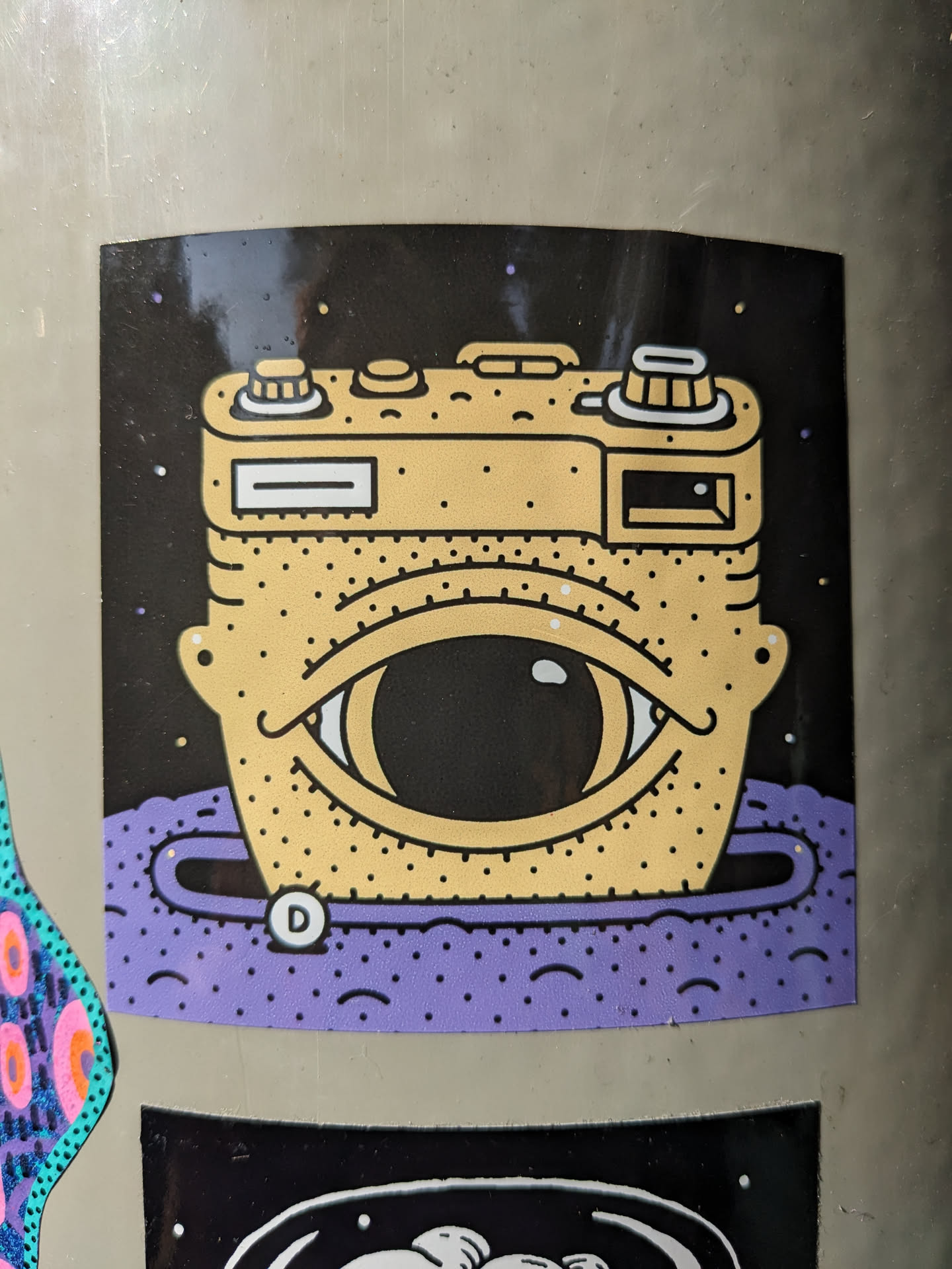Sticker of a camera with one eye, set in space. The camera is yellow and the background is black with purple accents. There is a small letter D in a circle on the purple part.