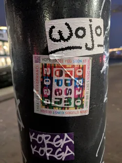 A sticker with the word "Wojo" written in a stylized font. Below it is a sticker advertising NDSM Open Studios with a QR code. There is another sticker at the bottom with "KORGA KORGA" written on it.