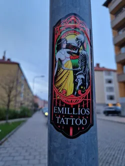 A sticker featuring an art deco style illustration of a woman dancing with a skeleton. The sticker is black and red with a yellow and green background. The text "Emilios Tattoo" is printed at the bottom.