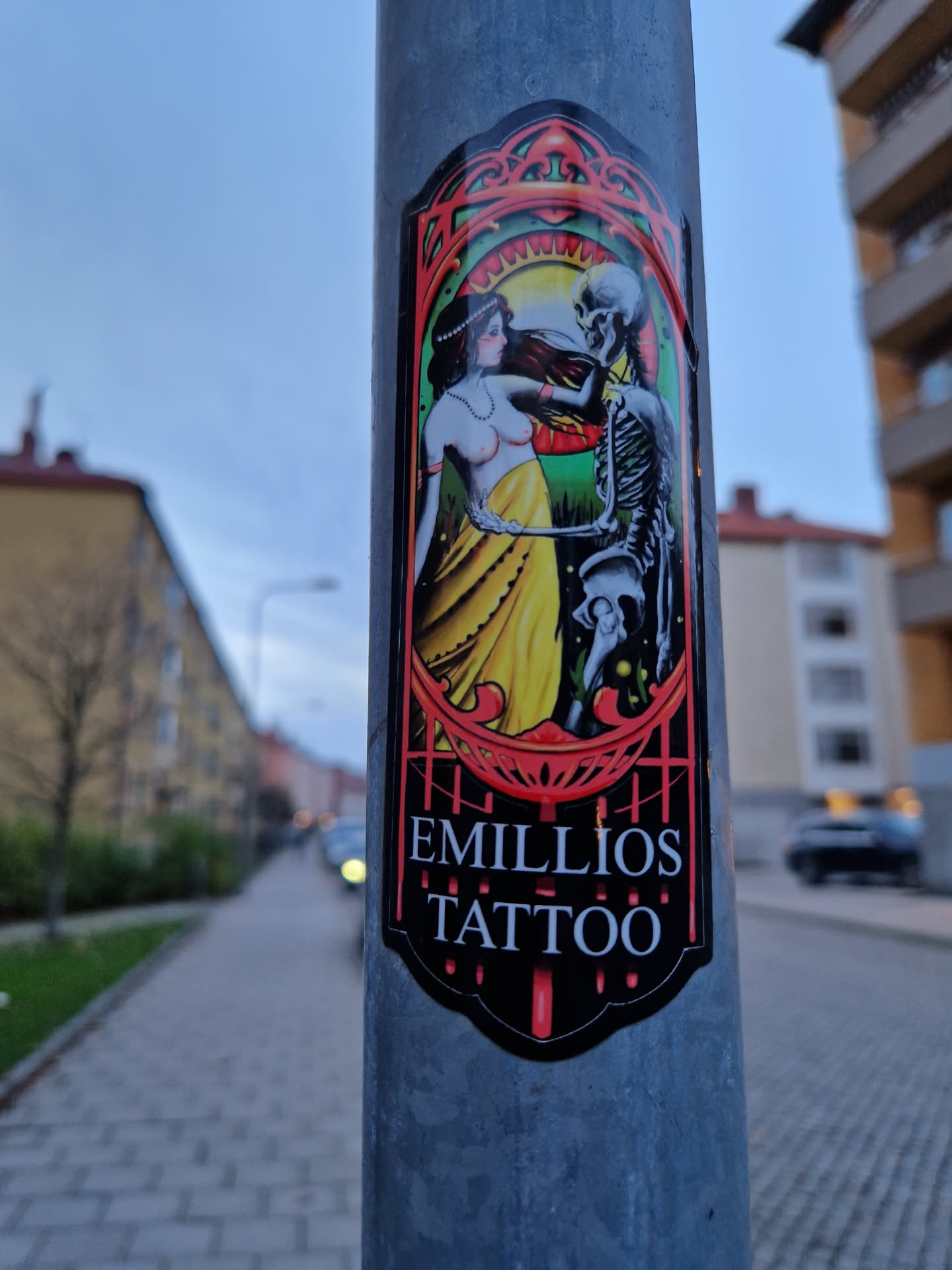 A sticker featuring an art deco style illustration of a woman dancing with a skeleton. The sticker is black and red with a yellow and green background. The text "Emilios Tattoo" is printed at the bottom.