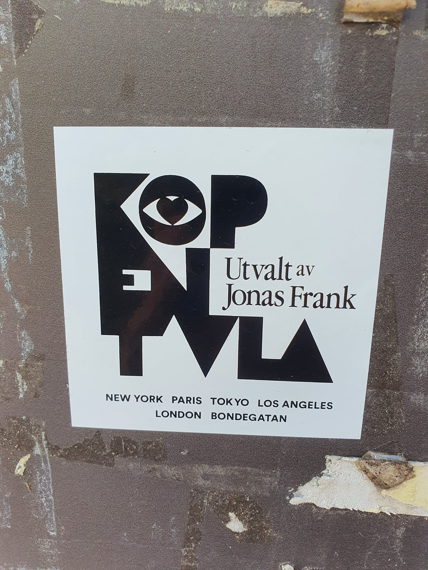Black and white sticker with the text "KOPENTVL" stylized with geometric shapes and an eye symbol with a heart in the middle. It also features the text "Utvalt av Jonas Frank" and a list of cities: New York, Paris, Tokyo, Los Angeles, London, and Bondgatan.