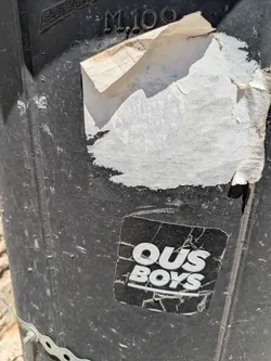 Close-up of a worn sticker with the text "OUS BOYS" in bold white letters. The sticker is on a black, textured surface, and the sticker itself is cracked and aged.