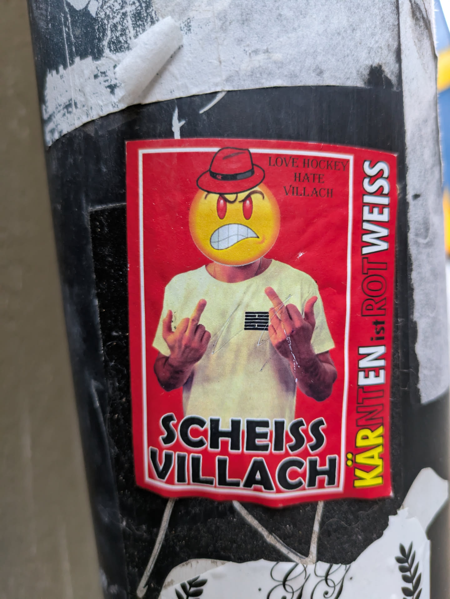Sticker featuring an angry emoticon wearing a red hat and giving the middle finger. The text on the sticker says "Love hockey hate Villach" and "Kärnten ist not Weiss" in German, which translates to "Carinthia is not white". There is also the phrase "Scheiss Villach", which means "Shit Villach".