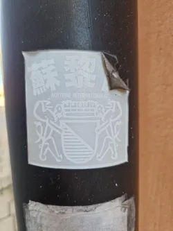 Close-up of a white Achtussig International sticker affixed to a dark pole. The sticker features a coat of arms flanked by two figures and Chinese characters at the top.