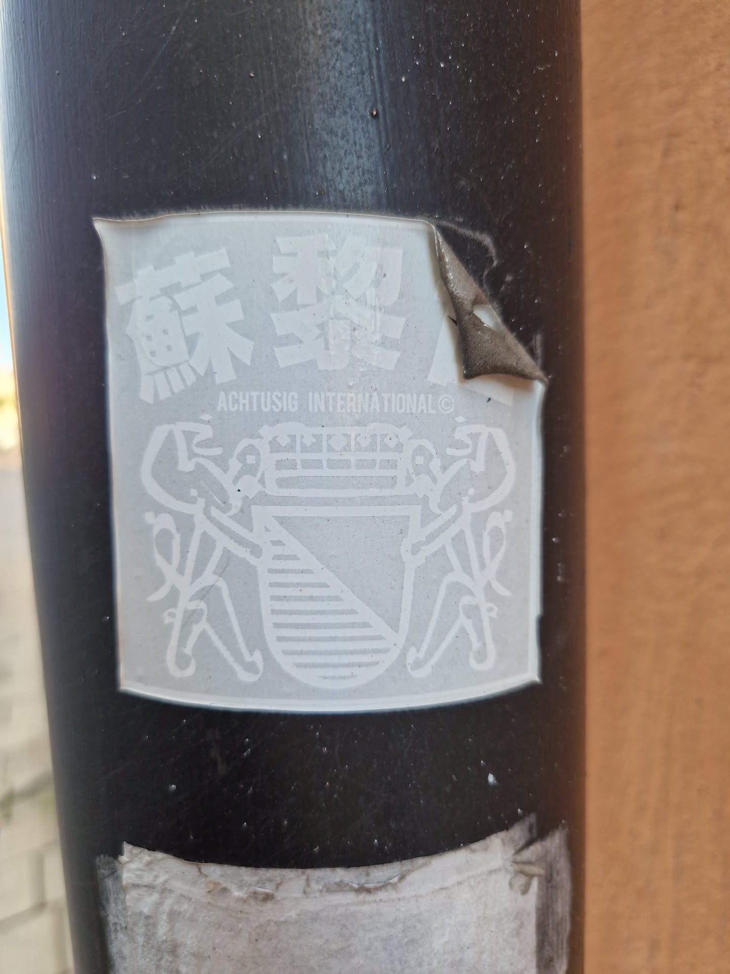 Close-up of a white Achtussig International sticker affixed to a dark pole. The sticker features a coat of arms flanked by two figures and Chinese characters at the top.