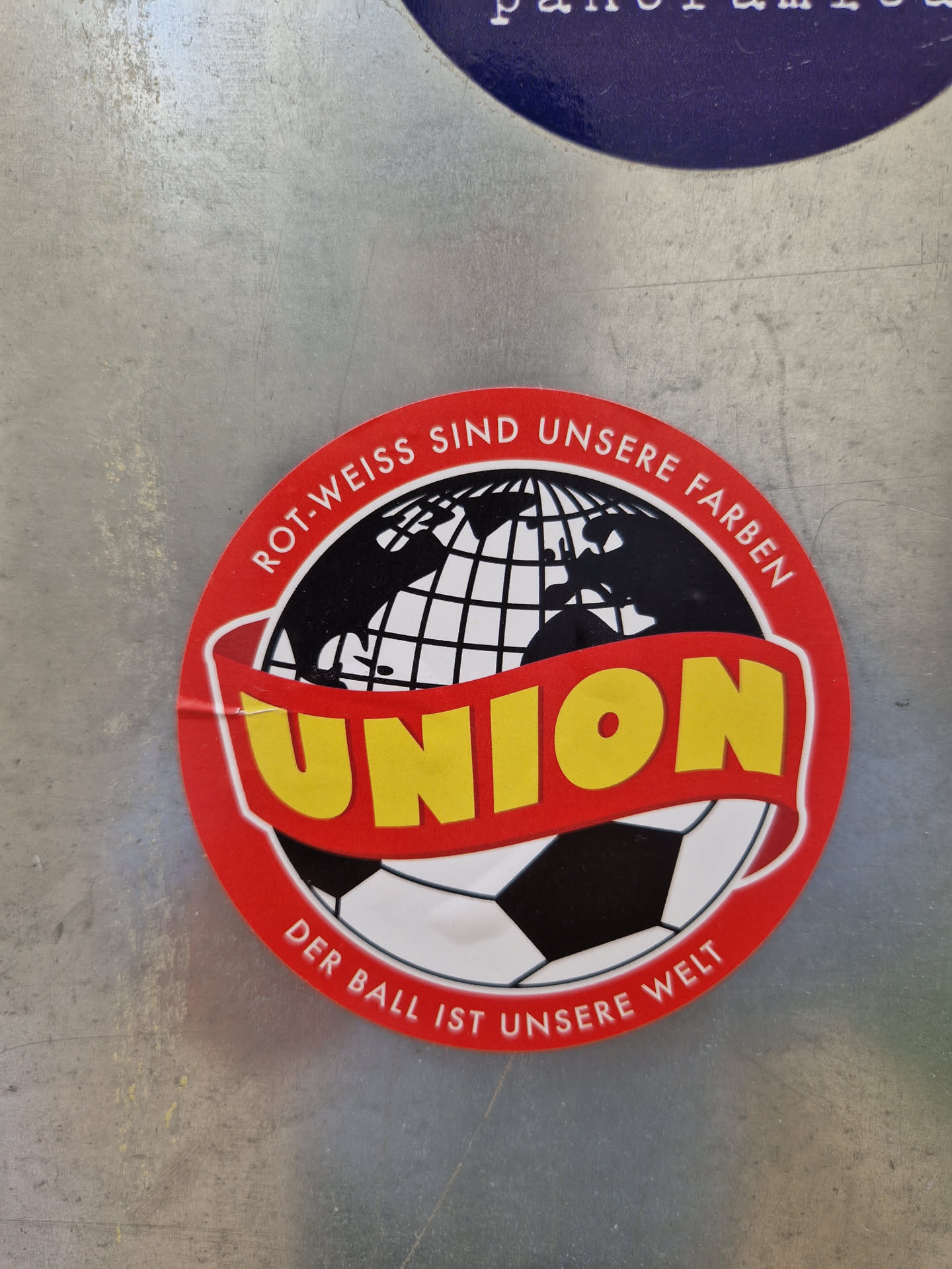 Round sticker with a red border and a globe in the center.  The word "UNION" is written in a yellow banner across the globe. A soccer ball is at the bottom. The text around the border says "Rot-Weiss sind unsere Farben" (Red-White are our colors) and "Der Ball ist unsere Welt" (The ball is our world) in German.