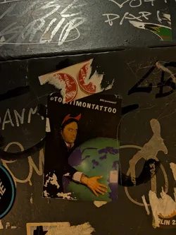 A vertical rectangular sticker set against a dark background, depicting a man wearing red devil horns and a dark suit. He stares intently forward while holding an inflatable globe representing Planet Earth in his arms. Across the top, white text displays the handle '@TOMTIMONTATTOO' with smaller text reading '80% permanenT' in the top right corner. The sticker exhibits weathering, torn corners, and surface abrasions.