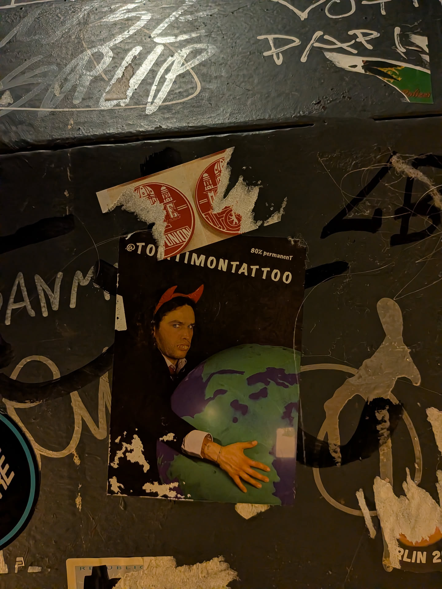 A vertical rectangular sticker set against a dark background, depicting a man wearing red devil horns and a dark suit. He stares intently forward while holding an inflatable globe representing Planet Earth in his arms. Across the top, white text displays the handle '@TOMTIMONTATTOO' with smaller text reading '80% permanenT' in the top right corner. The sticker exhibits weathering, torn corners, and surface abrasions.