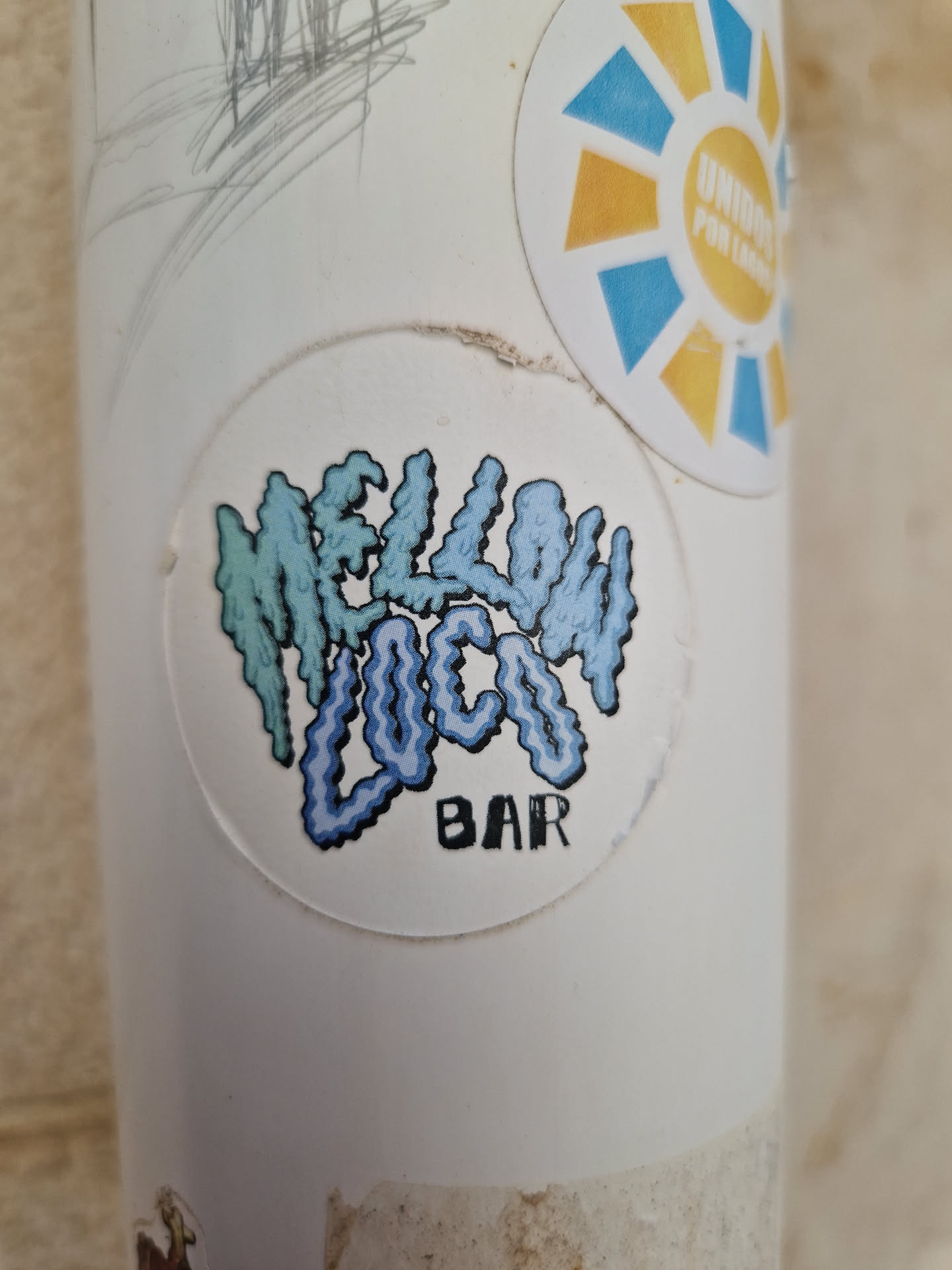 A sticker featuring the text "Mellow Loco Bar" in a stylized font. The text is in light blue and teal colors and has a pixelated or slightly distorted appearance. The sticker is circular and has a white background.