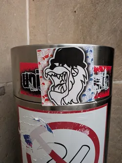 Street sticker A rectangular sticker placed on a metal surface, featuring a bold black-and-white graphic of a snarling, fanged creature or gorilla wearing a black bowler hat. The main figure is surrounded by a background pattern of smaller repeating sketches of the same character in red on the left side and blue on the right side.