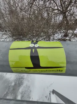 A lime green sticker with a black abstract figure resembling a stylized person or bird.  The SoundCloud URL soundcloud.com/raeubermukke is printed at the bottom. The sticker is affixed to a metal railing in a snowy outdoor setting.
