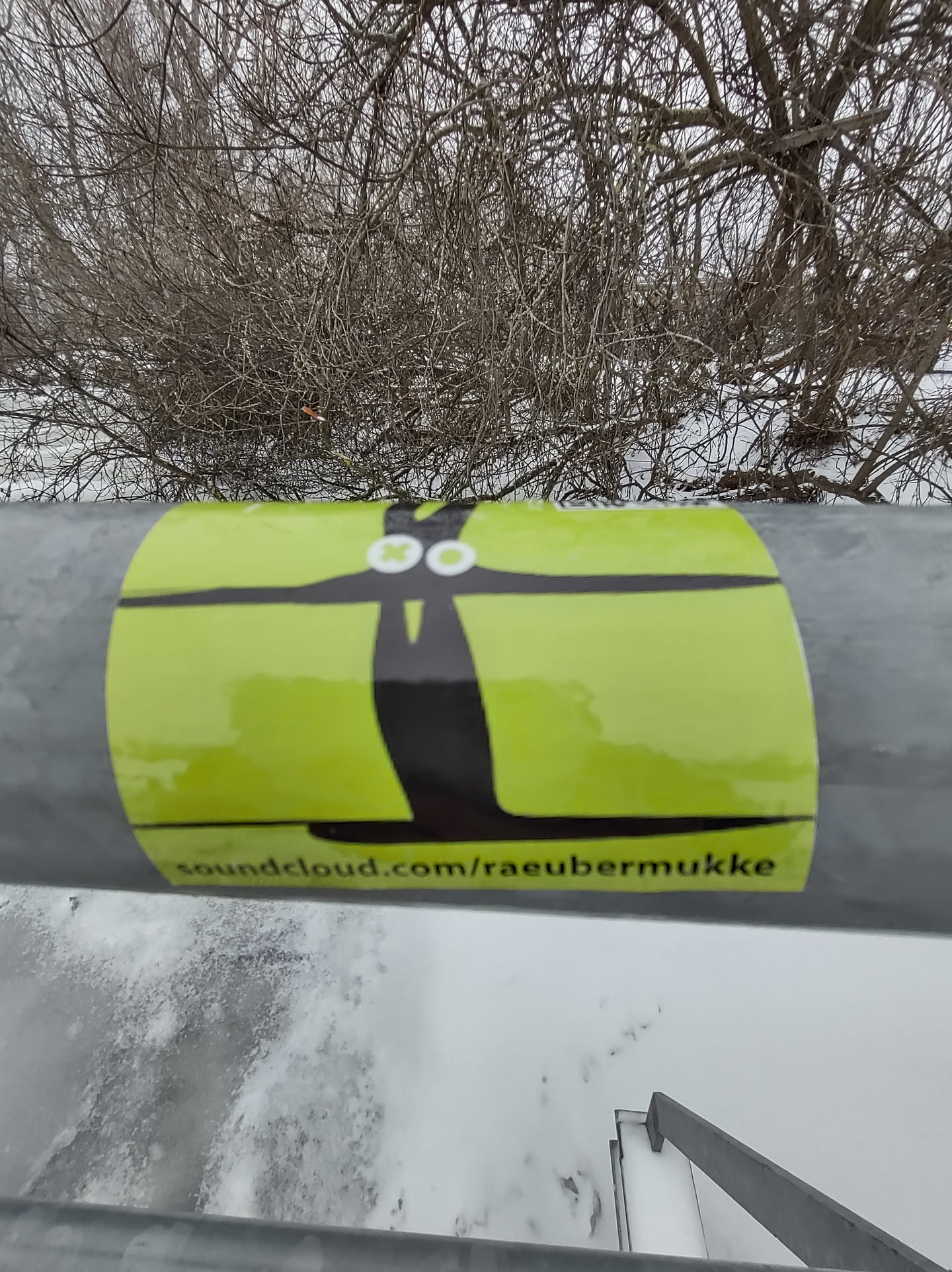 A lime green sticker with a black abstract figure resembling a stylized person or bird.  The SoundCloud URL soundcloud.com/raeubermukke is printed at the bottom. The sticker is affixed to a metal railing in a snowy outdoor setting.