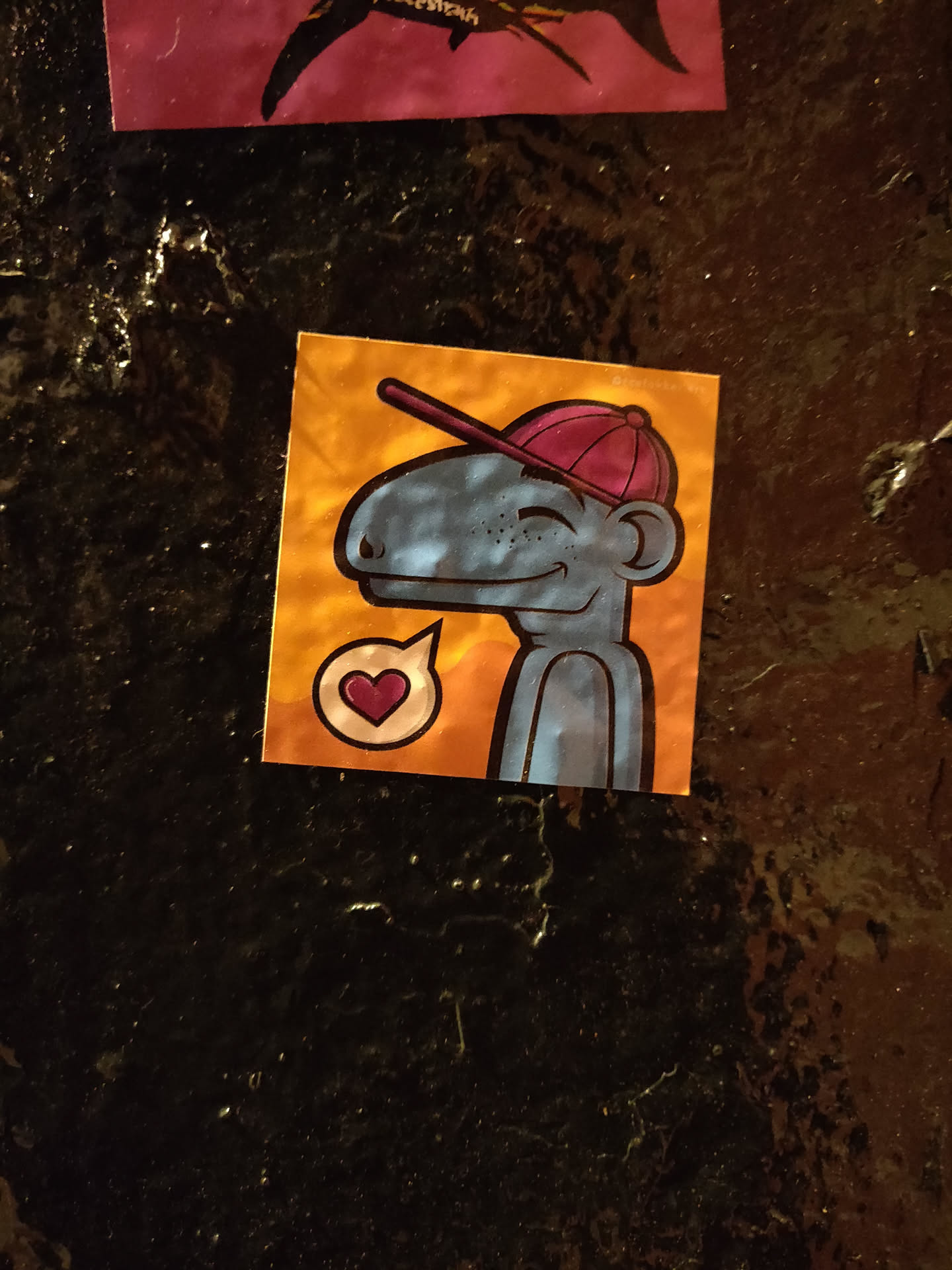 A small, blue creature wearing a red baseball cap is featured on this sticker. It has a friendly expression and a small heart-shaped speech bubble beside it, suggesting a message of love or affection. The sticker has a vibrant color scheme with an orange background.