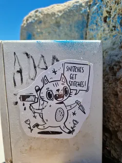 A black and white sticker featuring a cartoon cat holding a paint roller and a knife. The cat's speech bubble says: "Snitch get stitches!"