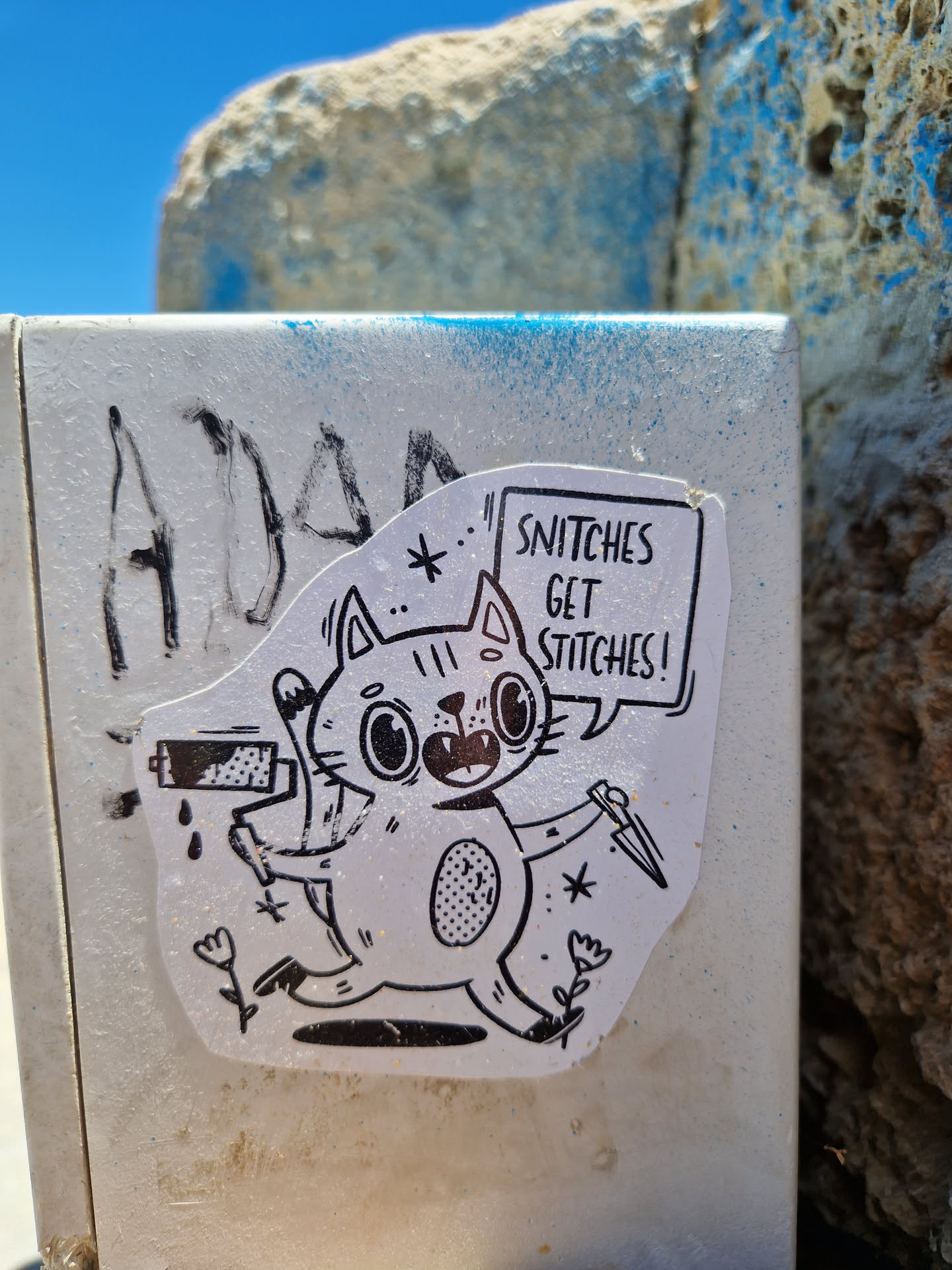 A black and white sticker featuring a cartoon cat holding a paint roller and a knife. The cat's speech bubble says: "Snitch get stitches!"