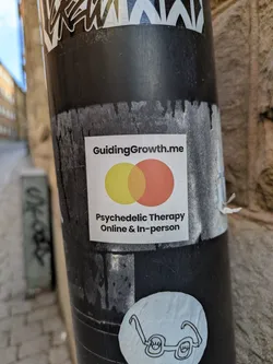 A square sticker on a black post features the text "GuidingGrowth.me" at the top, followed by two overlapping circles in yellow and orange. The text "Psychedelic Therapy Online & In-person" is printed below the circles. The sticker advertises a therapy service.