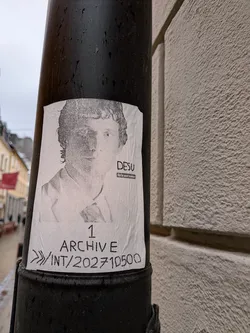 The sticker is a rectangular, white paper print affixed to a dark, textured pole. It features a grainy, black-and-white portrait of a man with curly hair, looking forward. His left side is partially obscured by shadow. He appears to be wearing a collared shirt and a jacket. Below the portrait, there is text in a handwritten-style font. The paper shows signs of wear, including wrinkles and a slightly curled bottom edge, indicating it's a real-world paste-up or sticker.