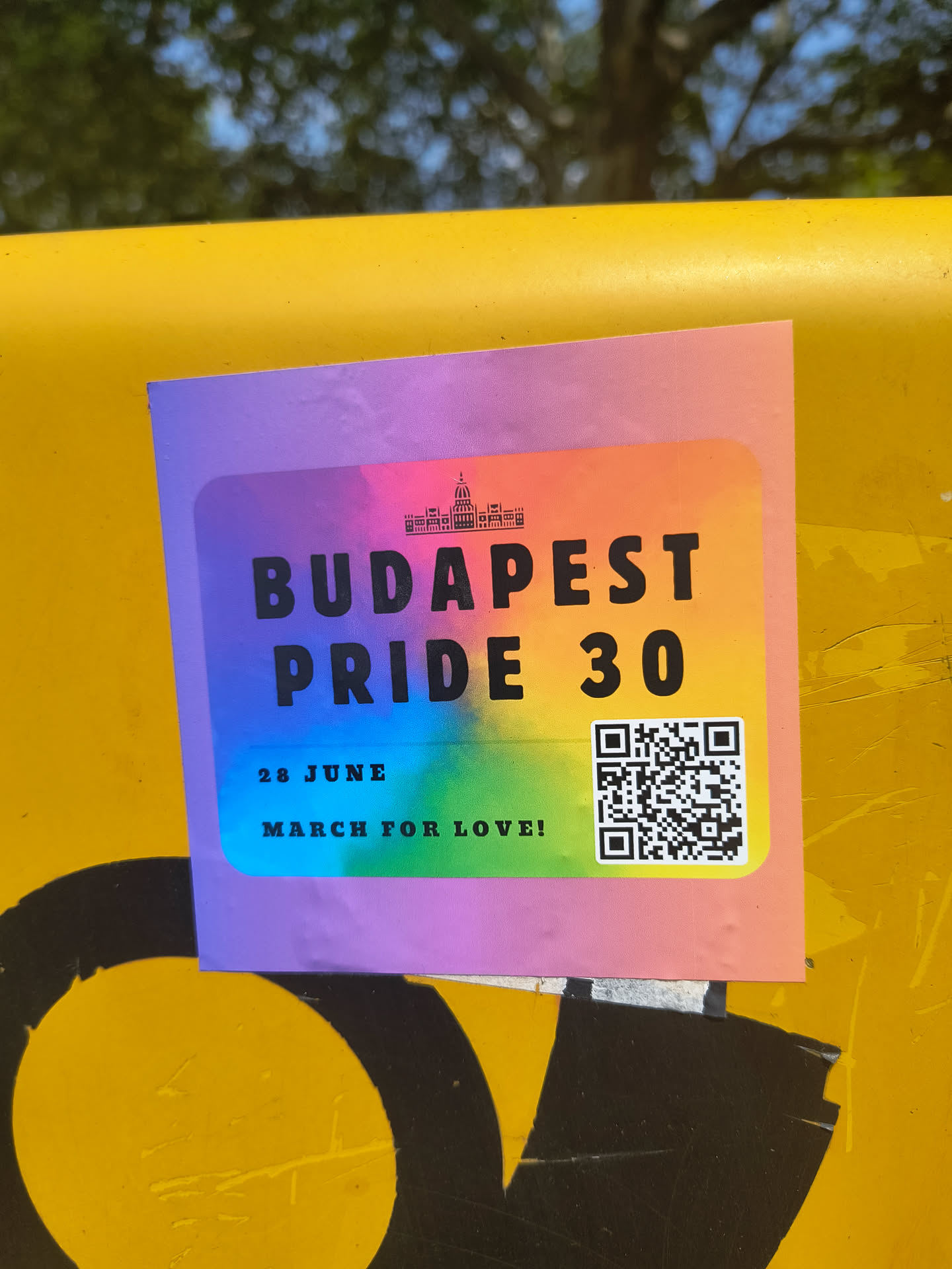 A rectangular sticker with rounded corners, featuring a bold rainbow gradient background transitioning from purple, blue, green, yellow, orange, to pink. At the top center, there's a black silhouette of a prominent building, likely the Hungarian Parliament. The main text 'BUDAPEST PRIDE 30' is printed in large, black, sans-serif font across the center. Below this, in smaller black text, it reads '28 JUNE' and 'MARCH FOR LOVE!'. A black and white QR code is prominently displayed in the bottom right corner of the sticker. The sticker appears to be adhered to a textured yellow surface, showing slight wear and adherence characteristics.