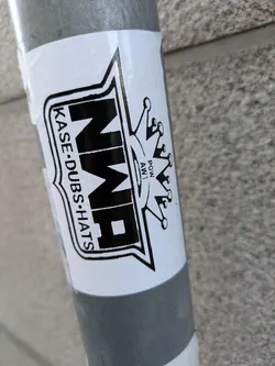 A black and white sticker featuring the NWA logo and the text "KASE-DUBSHATS". A crown is depicted next to the logo.  The sticker is affixed to a gray metal pole.