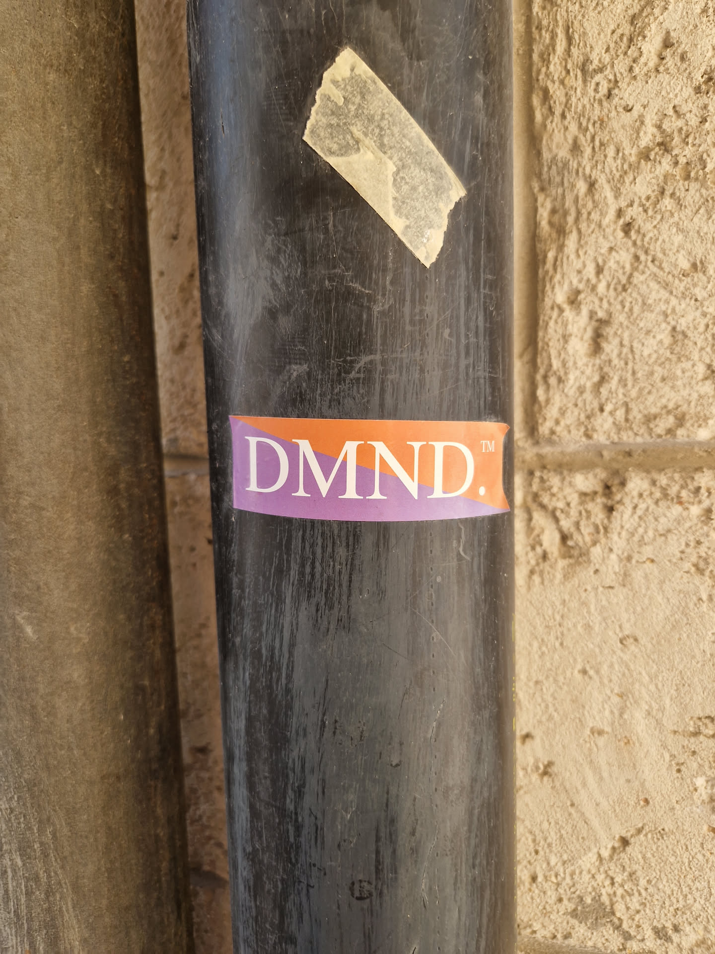 A close-up shot of a DMND. sticker affixed to a dark-colored pipe. The sticker is rectangular with an orange background and purple text.  The pipe is weathered and shows signs of wear.