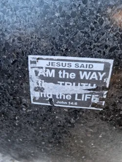 A worn sticker with a biblical quote from John 14:6. The sticker is affixed to a dark gray, speckled surface, possibly asphalt or concrete.
