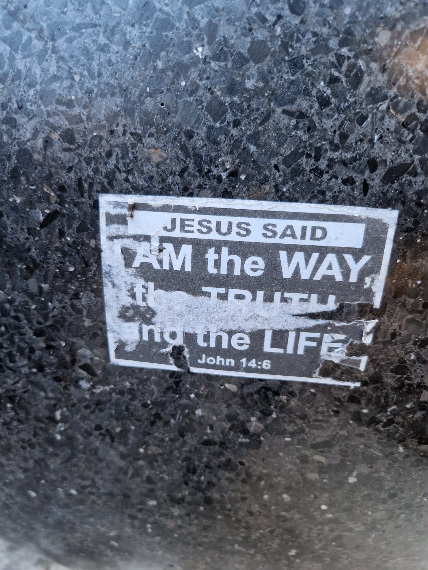 A worn sticker with a biblical quote from John 14:6. The sticker is affixed to a dark gray, speckled surface, possibly asphalt or concrete.