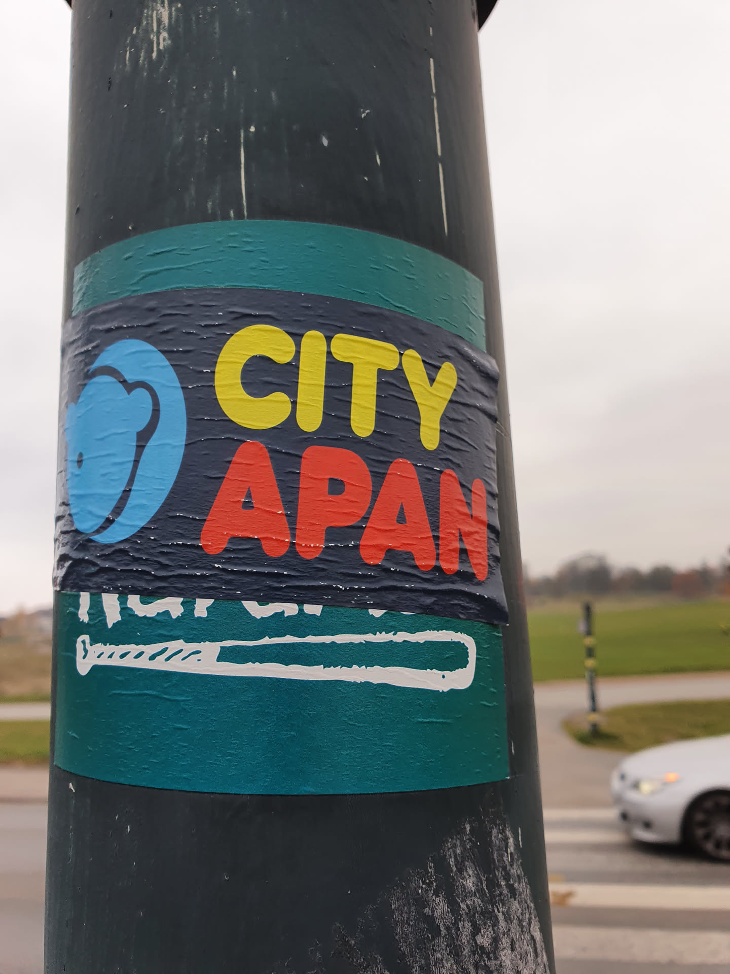A teal sticker featuring the words 'CITY APAN' in yellow and red lettering. There is also a small graphic of what looks like a baseball bat at the bottom and a circular logo in blue with an indiscernible symbol to the left of 'CITY'. The sticker appears to be affixed to a dark-colored pole outdoors.