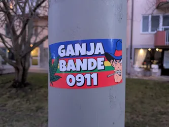 A sticker featuring a cartoon character with a cowboy hat smoking a cigarette, along with the text "GANJA BANDE 0911" and a marijuana leaf. The sticker is affixed to a pole outdoors.