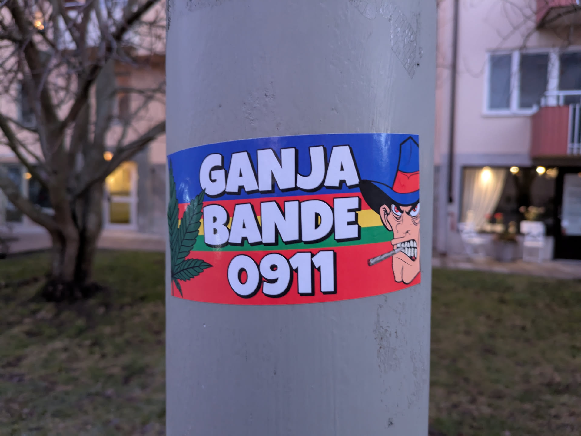 A sticker featuring a cartoon character with a cowboy hat smoking a cigarette, along with the text "GANJA BANDE 0911" and a marijuana leaf. The sticker is affixed to a pole outdoors.